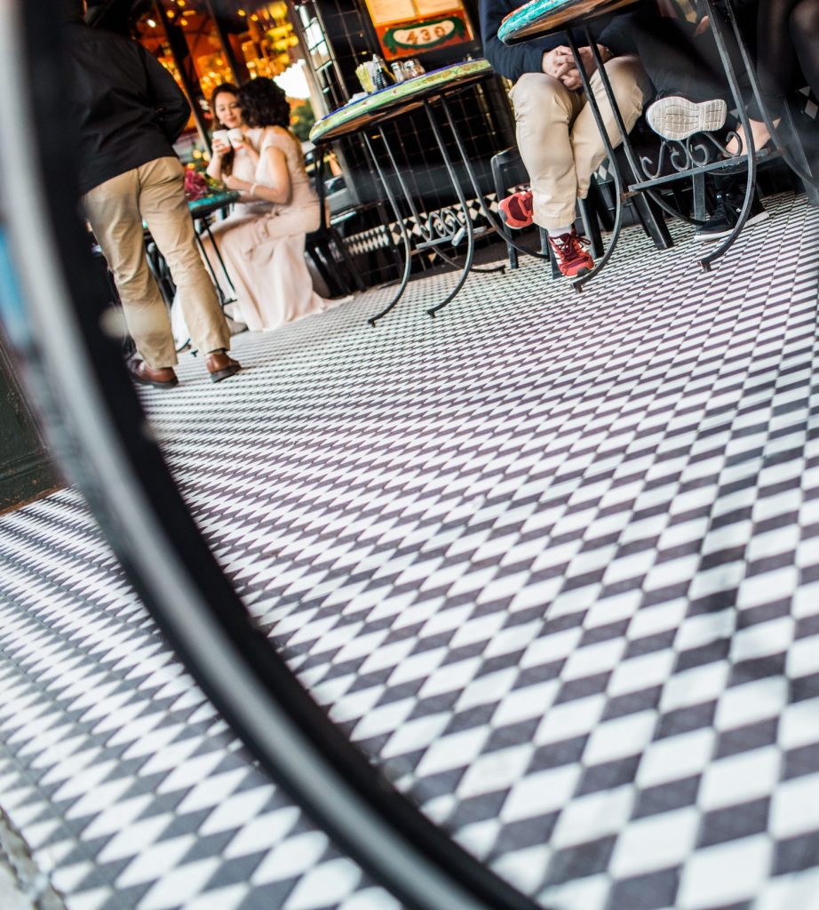 Two women at a table in the background clink coffee cups on a foreground of black and white sidewalk checker tiles.